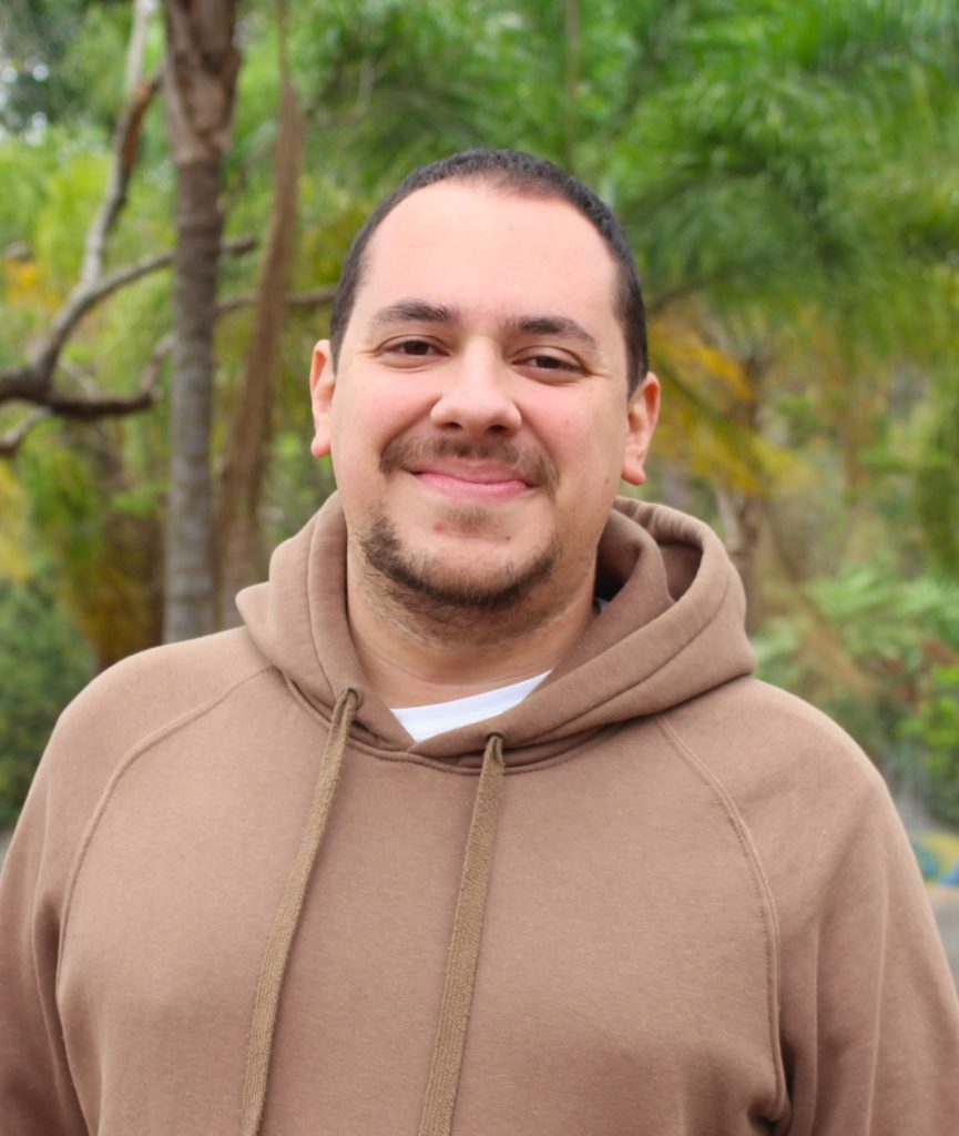 Heitor sorri para a câmera em frente a árvores. Ele tem cabelo ralo castanho, uma cavanhaque também castanho e pele clara. Veste um moletom de cor creme.