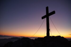 Uma cruz levantada por cabos está no topo de uma montanha, com um pôr do sol ao fundo