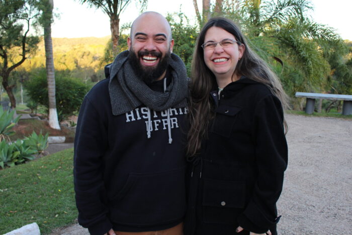 Um homem branco careca com barba sorri está ao lado de uma mulher branca com cabelos castanhos compridos. Ambos sorriem para a foto. Ao fundo, vê se algumas plantas.