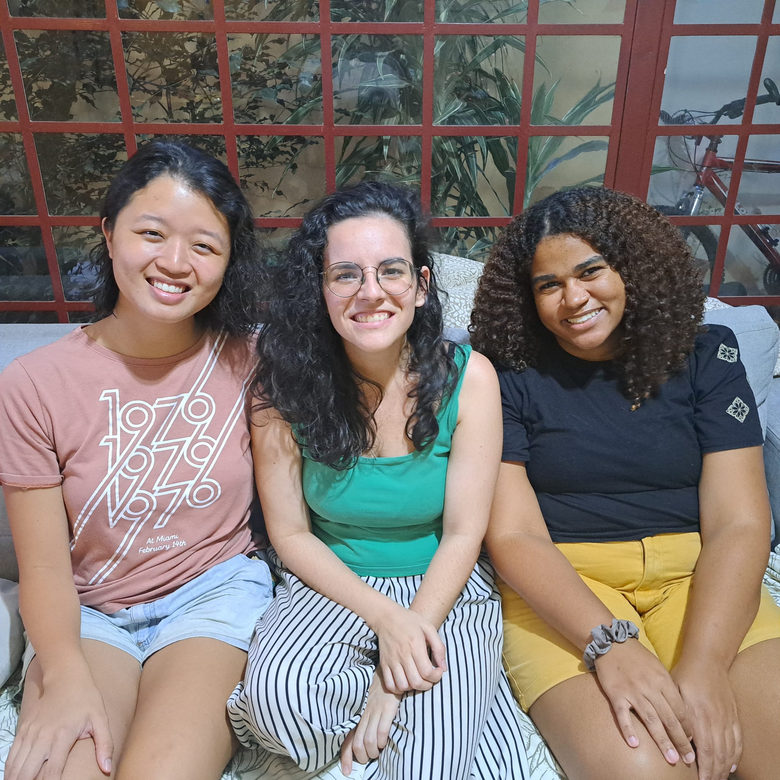 Três meninas sentadas, sorriem para a foto. São as participantes do Siga-me 2023, olhando para a câmera. Elas vestem roupas informais e coloridas. A primeira é asiática, a segunda branca e a terceira negra.