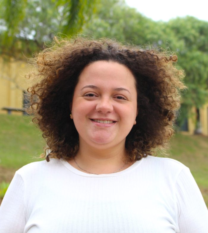 gabriela Assessora Gabriela sorri para a câmera. Ela tem cabelos castanhos cacheados de tamanho curto, pele clara e veste uma blusa branca. No fundo, vê-se árvores e um prédio amarelo.