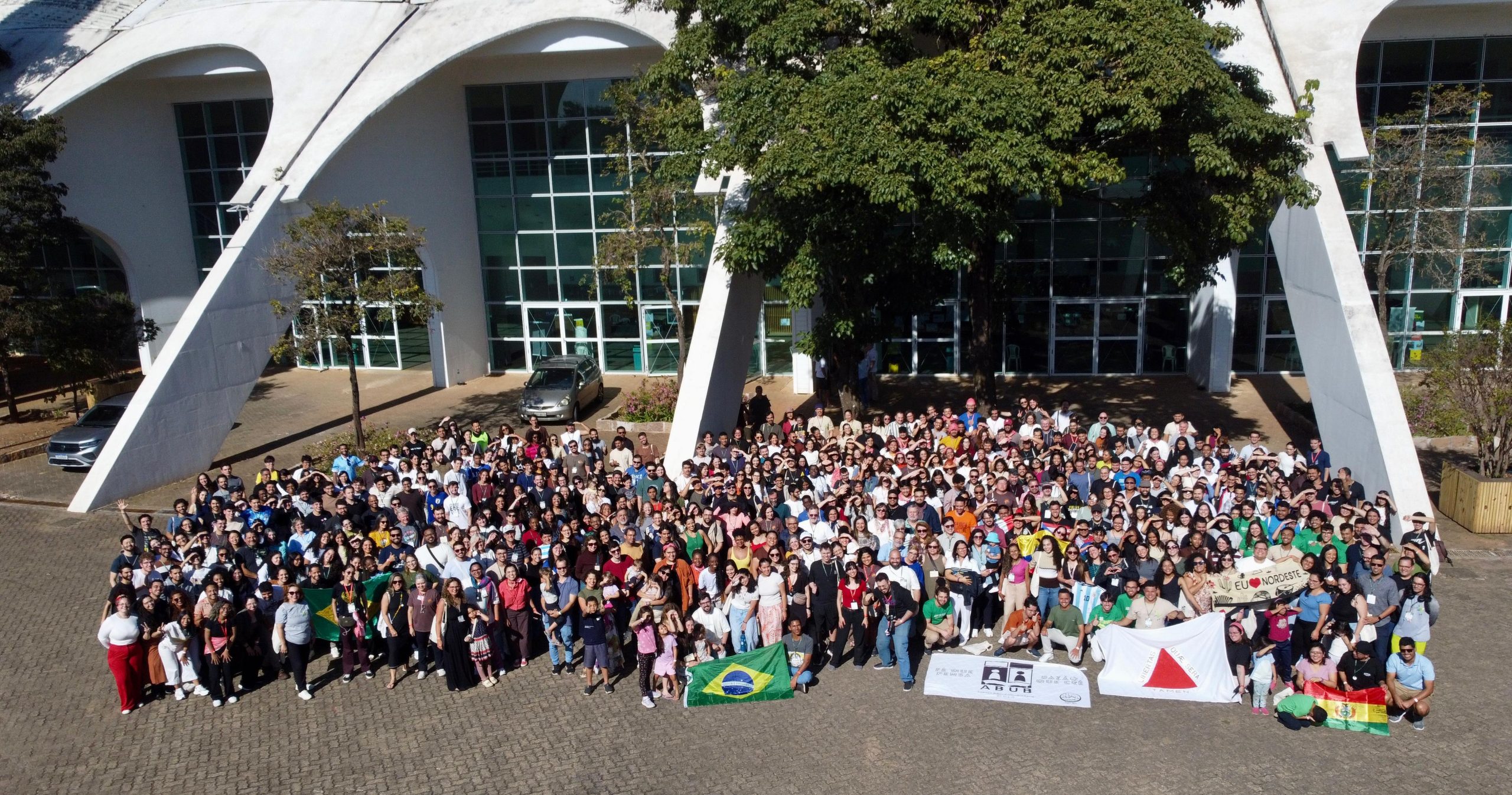 Grupo grande de pessoas com roupas coloridas posam para foto oficial do congresso Missão 2026: Amanhar em frente a um prédio branco e árvores grandes durante o dia. A foto está de cima para baixo e é possível ver bandeiras com logo da ABUB, bandeira do Brasil, de Minas Gerais e outros países da América Latina.